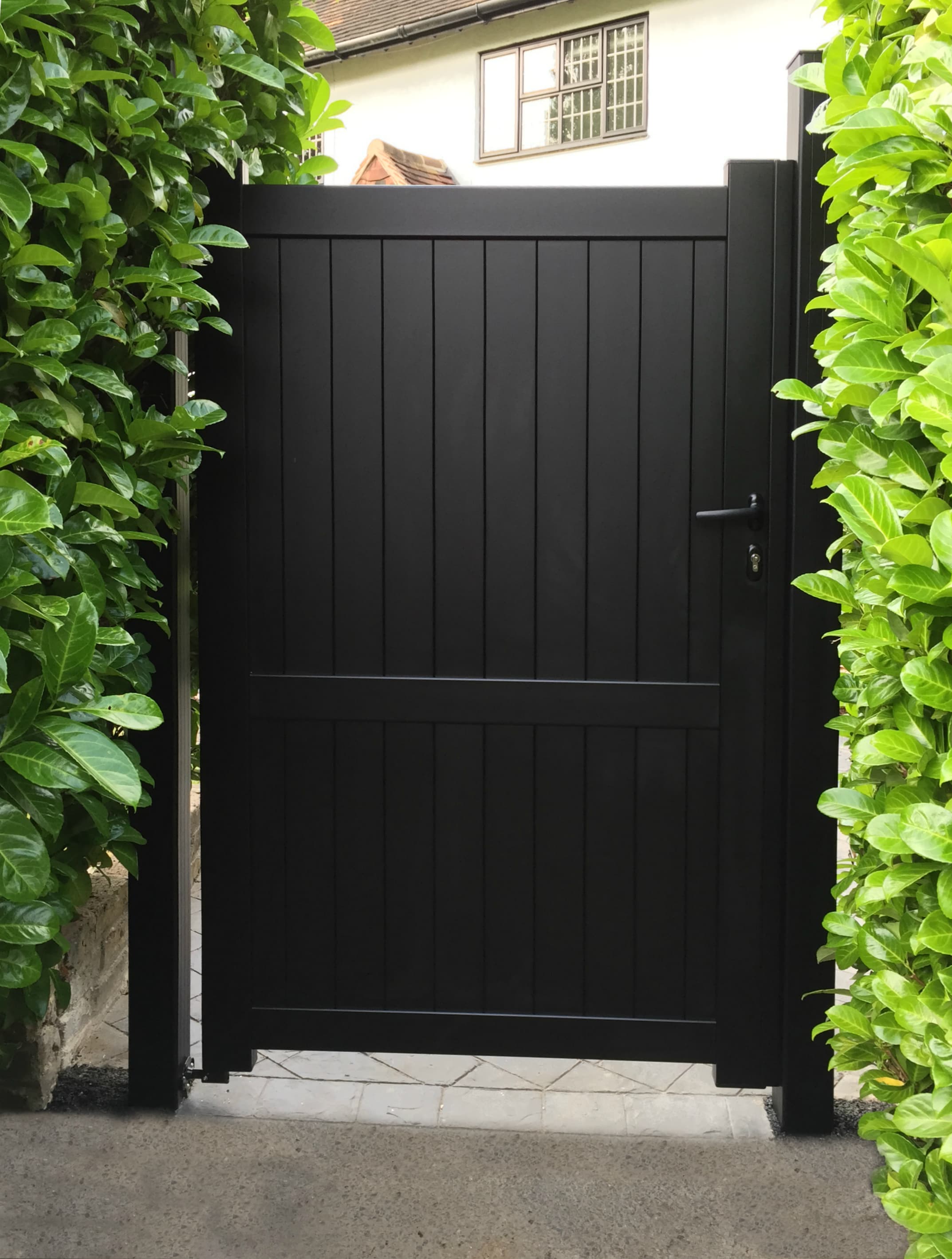 Dark black wooden gate framed by lush green plants, with a glimpse of a traditional house in the background featuring white windows.